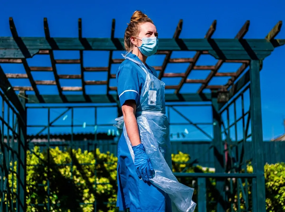 Female healthcare worker in blue PPE, mask, gloves, and plastic apron stands outdoors under a wooden pergola against a bright blue sky.
