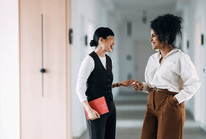 Two women coworkers in a bright office hallway, smiling as they chat; one holds a red folder.