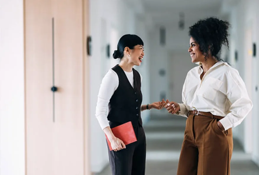 Two women coworkers in a bright office hallway, smiling as they chat; one holds a red folder.